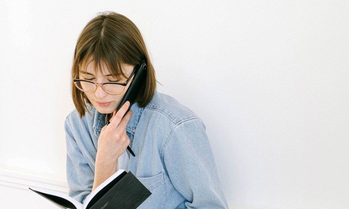 A woman writing notes by hand while taking a call on a desk phone.